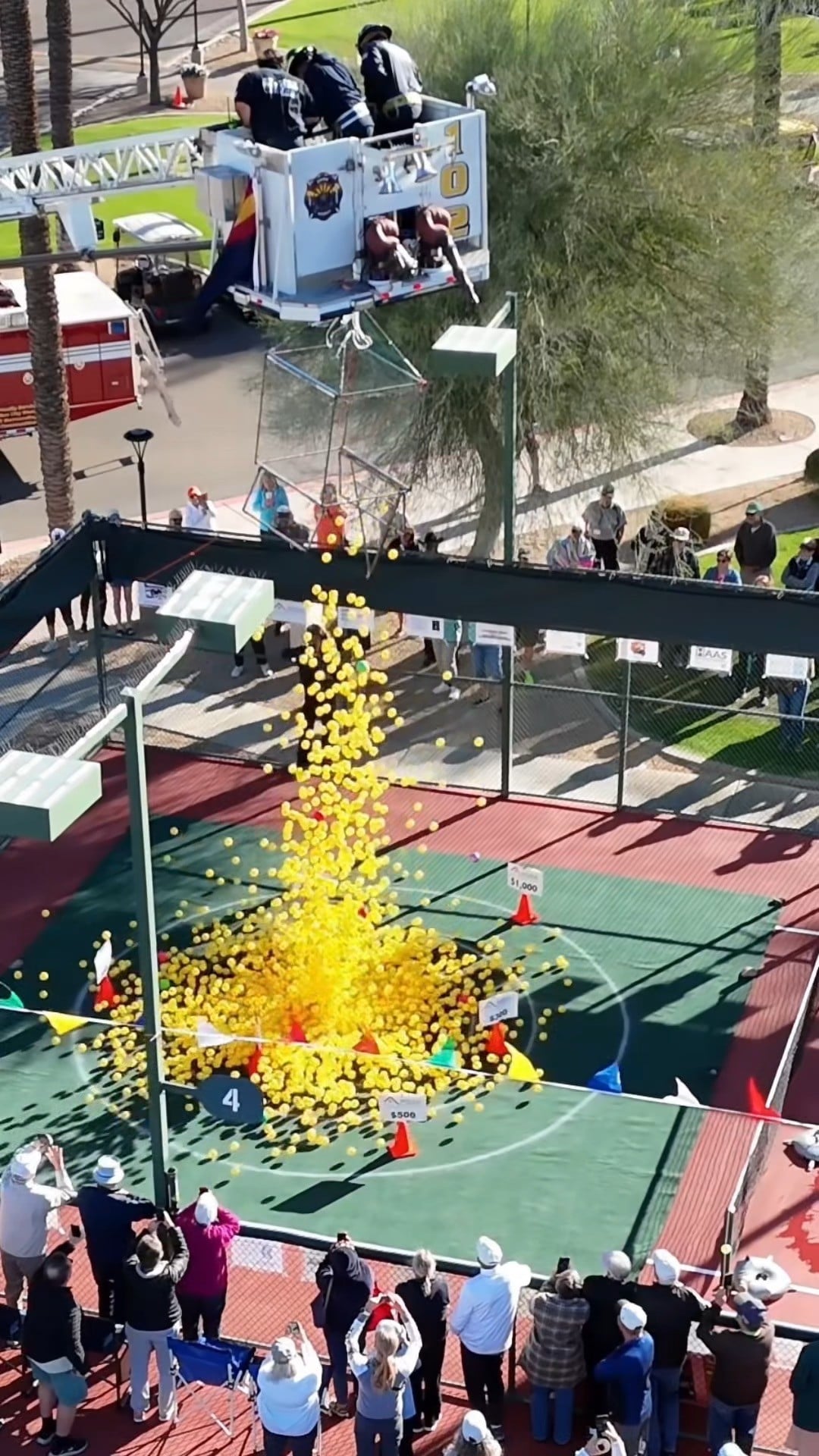 Firefighters release hundreds of bright yellow pickleballs from a ladder truck onto the Palm Ridge Pickleball Center court during the Sun City West Pickleball Ball Drop fundraiser, as spectators watch and cheer from the sidelines.