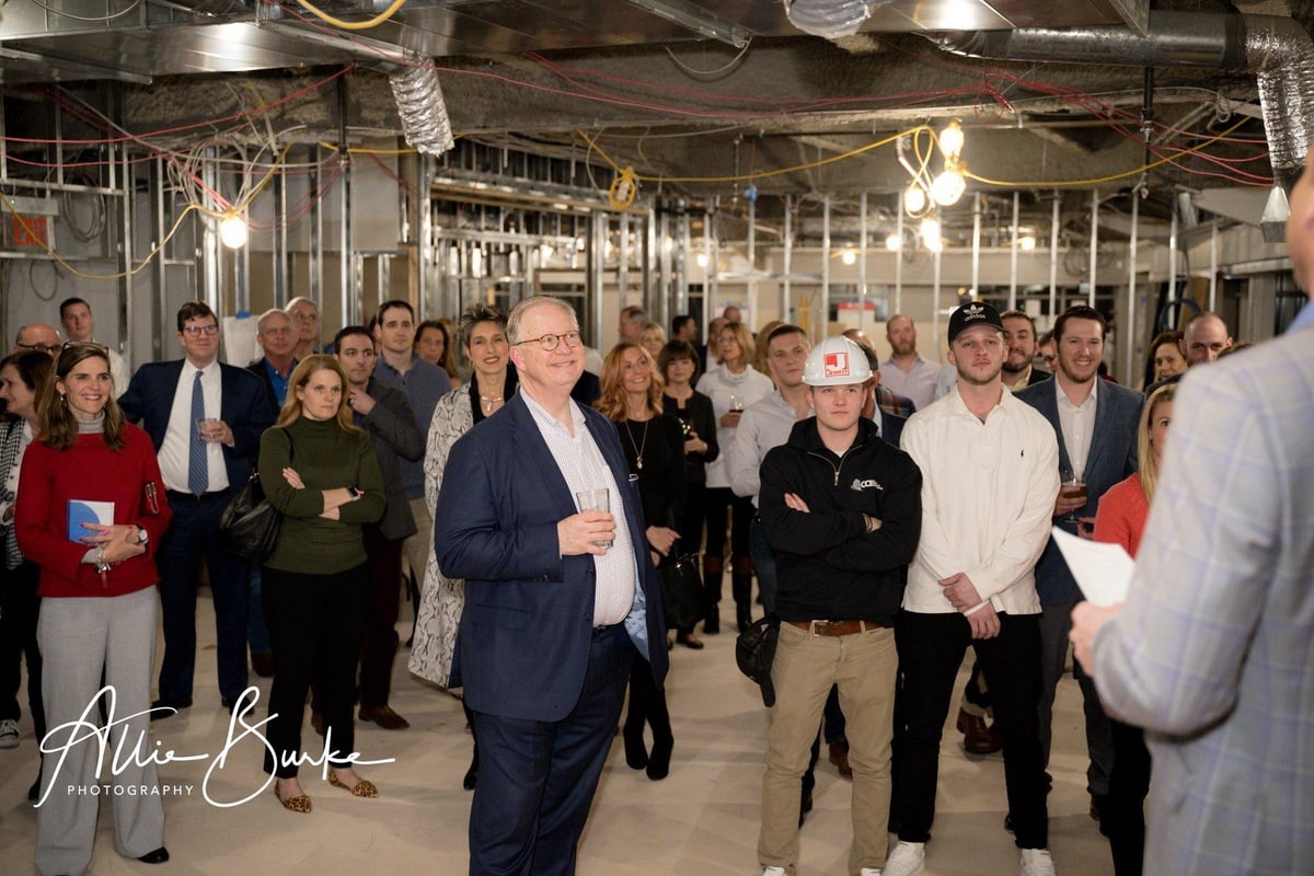 Crowd of guests gathered at a Jewett Construction event inside a building under construction, with exposed ceilings and wires overhead.
