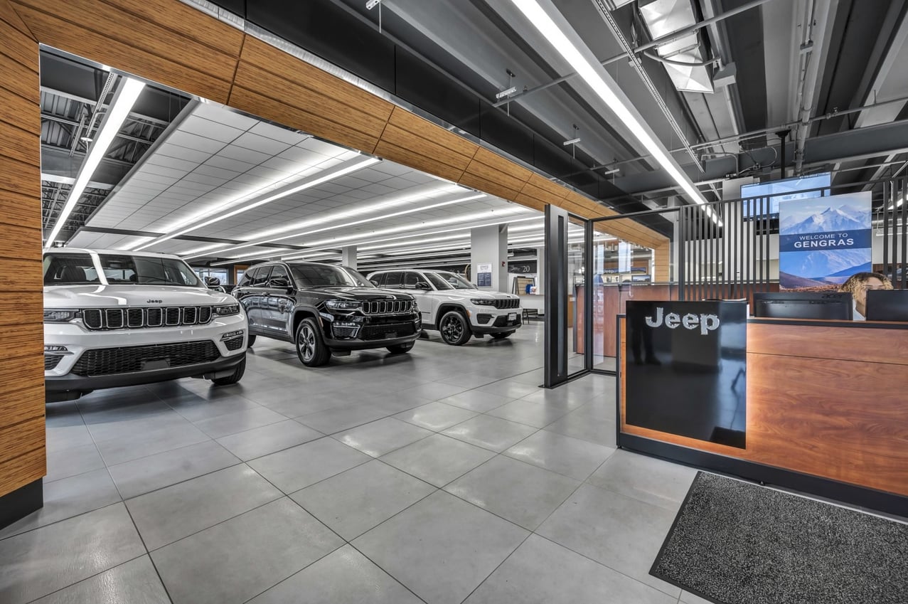 Modern Jeep dealership showroom featuring white and black Jeep SUVs parked in a polished, well-lit space with sleek architectural finishes. A reception desk labeled “Jeep” and a welcome sign for Gengras are visible on the right.