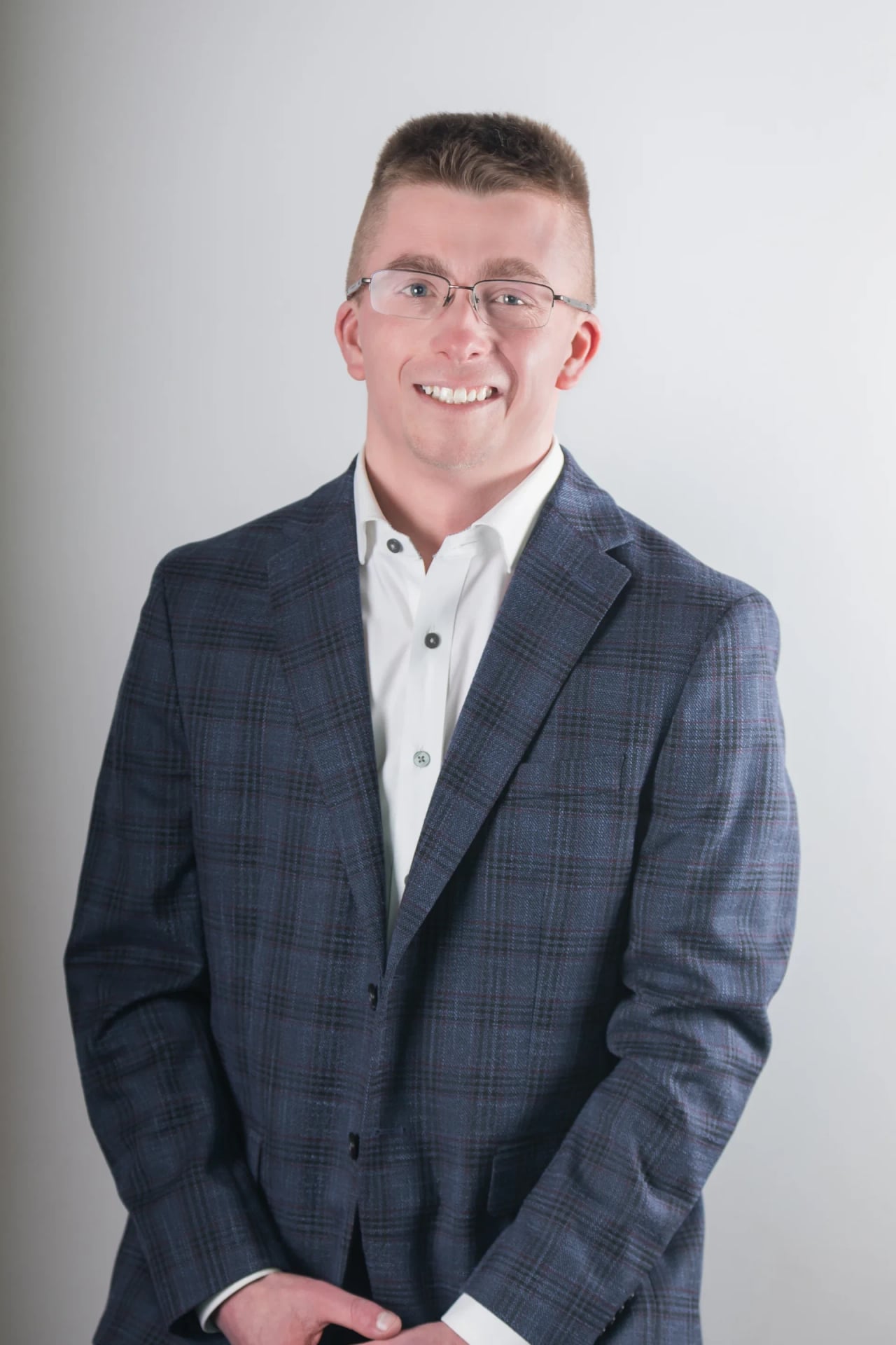 Professional portrait of a smiling man wearing glasses, a dark blue plaid blazer, and a white dress shirt, standing against a light gray background.