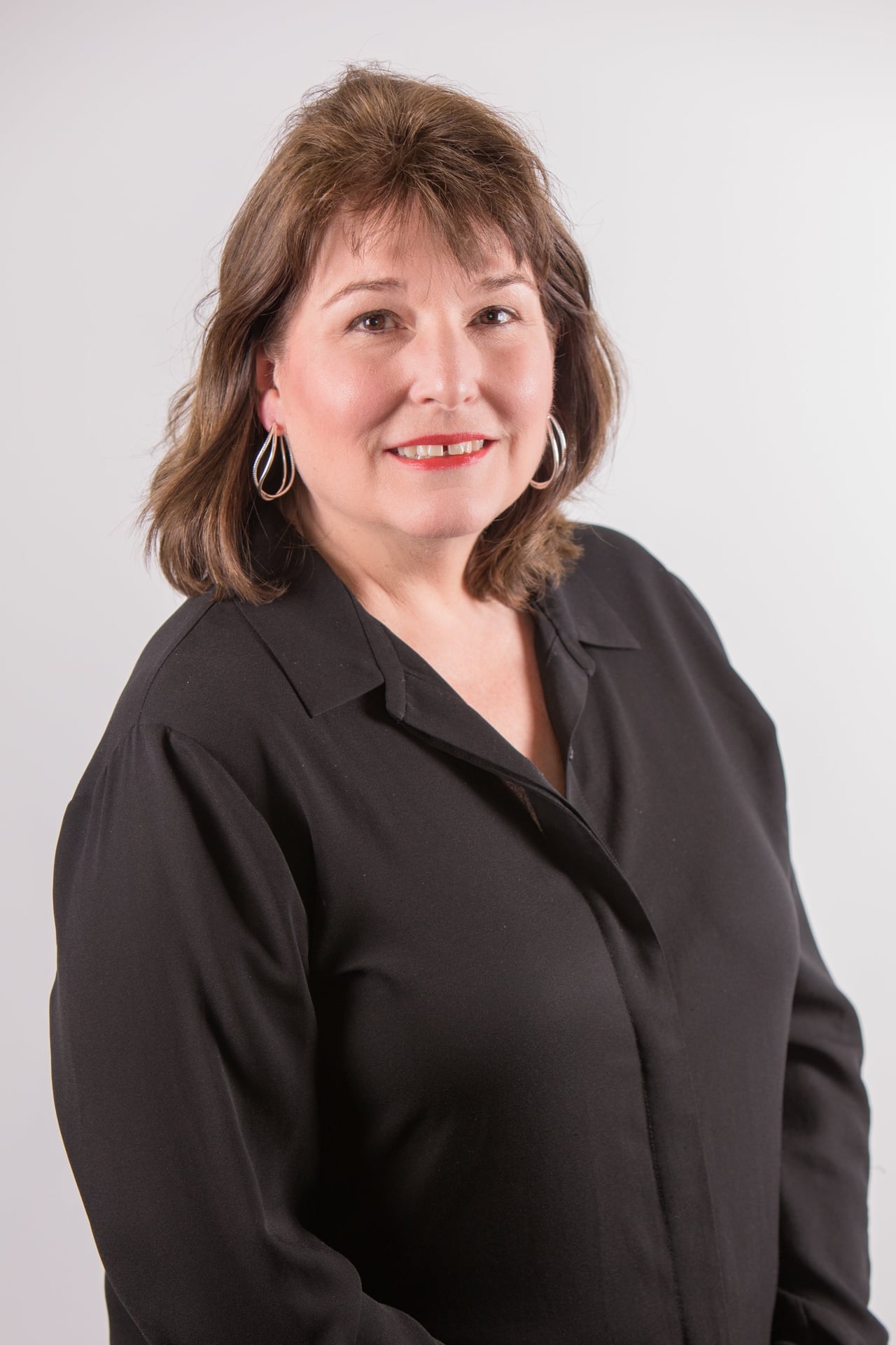 Professional portrait of a woman with shoulder-length brown hair wearing hoop earrings and a black blouse, smiling against a light gray background.