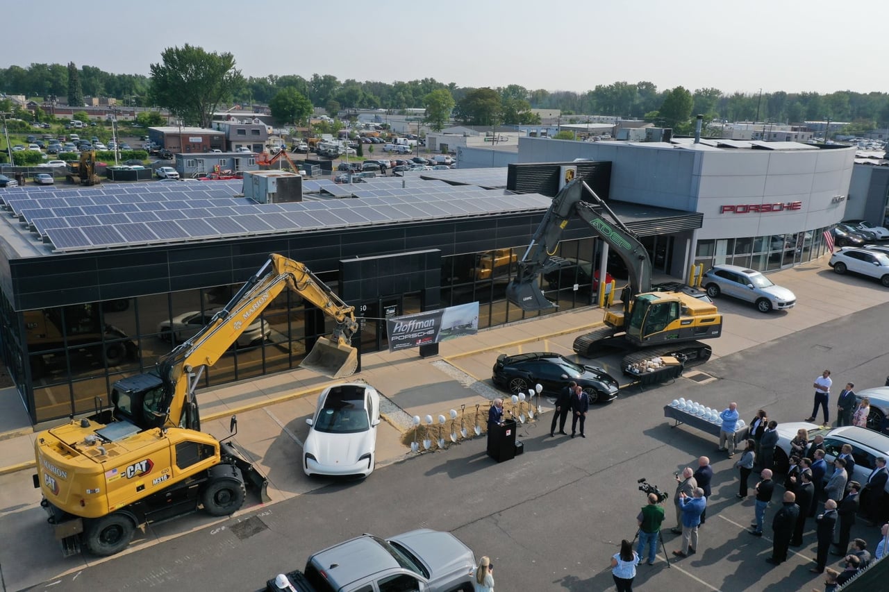 Aerial view of a groundbreaking ceremony at Hoffman Porsche, featuring construction equipment, solar panels on the dealership roof, white and black Porsche vehicles, a podium with speakers, and a crowd of attendees gathered for the event.