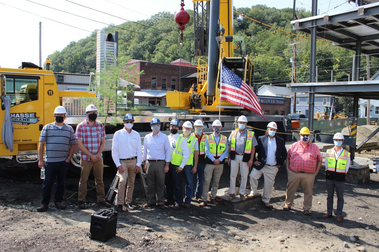 A group of construction professionals in hard hats and safety vests standing in front of a crane holding a steel beam adorned with an American flag and a tree, at a construction site during a topping-out ceremony.