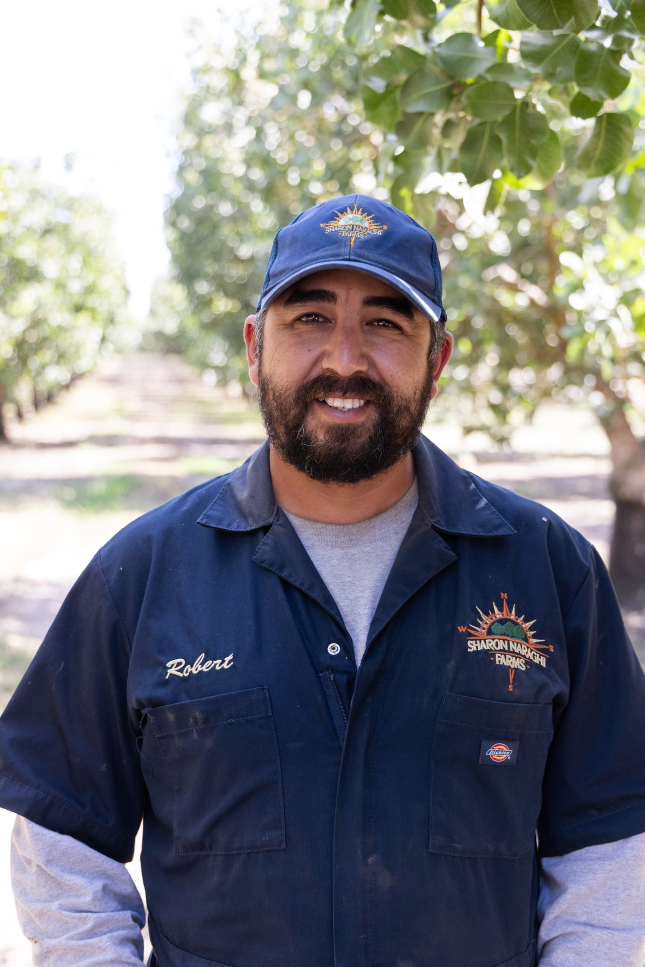 An adult in a navy work shirt and cap stands centered in an orchard row, smiling. Soft background blur isolates the subject, representing field leadership that coordinates irrigation, equipment, and harvest timing to protect crop quality.