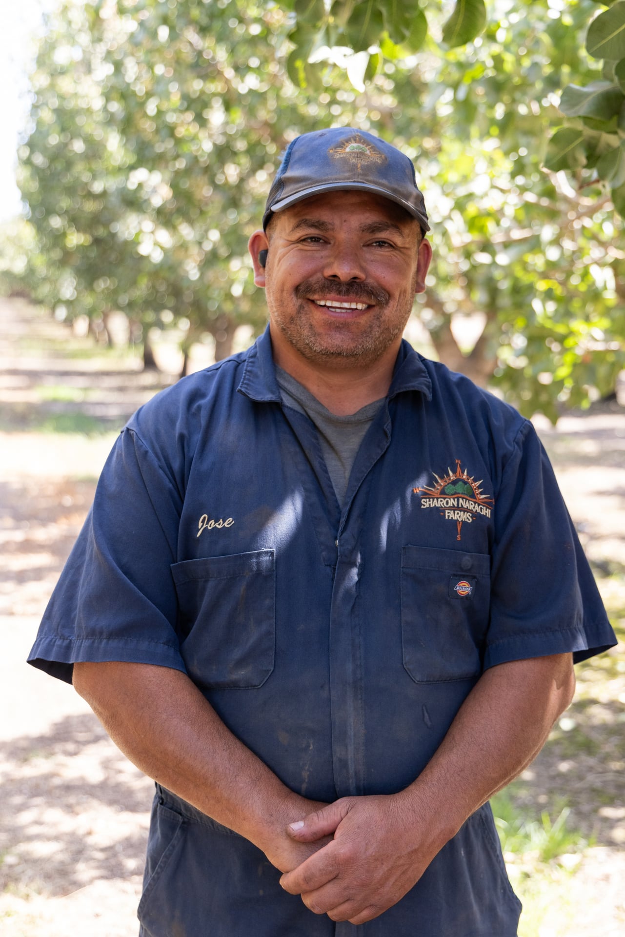 An adult in a navy shop uniform and cap stands with hands clasped in a tree lane. The portrait highlights the skilled service that keeps shakers, sweepers, and conveyors reliable—critical to clean, timely harvests for Central Valley growers.