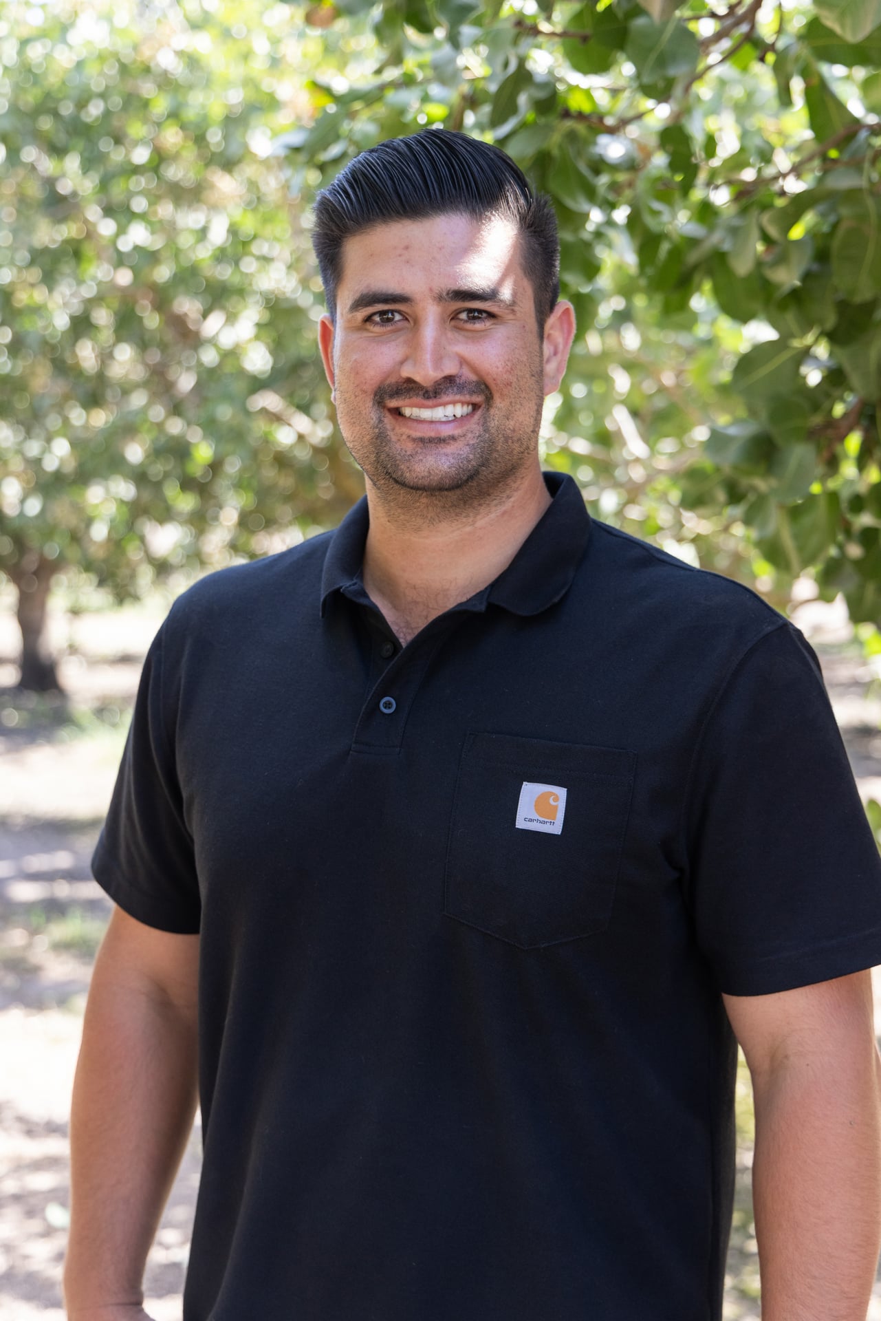A single adult in a polo stands mid-row in the orchard, smiling toward camera. The clean, straightforward framing communicates professionalism and day-to-day operational oversight within Sharon Naraghi Farms’ harvesting and crop programs.