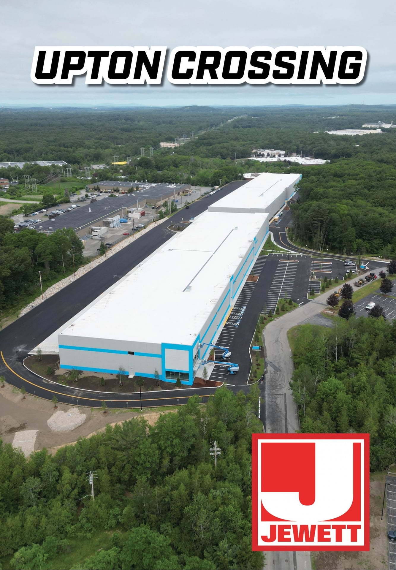 Aerial view of the newly completed Upton Crossing industrial building surrounded by forest and parking areas, with bold text “Upton Crossing” at the top and the Jewett Construction logo at the bottom right.