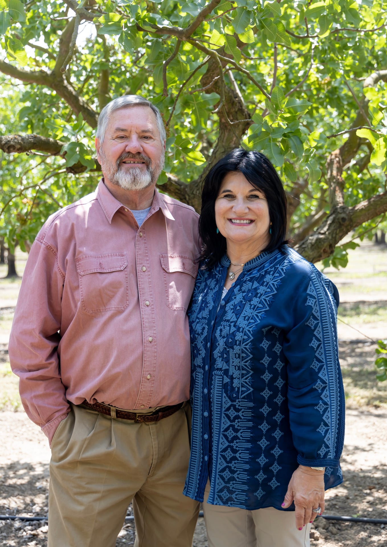 Two adults stand together beneath mature trees, smiling in soft shade. The portrait conveys family ownership and hands-on leadership central to the farm’s multigenerational legacy and commitment to quality.