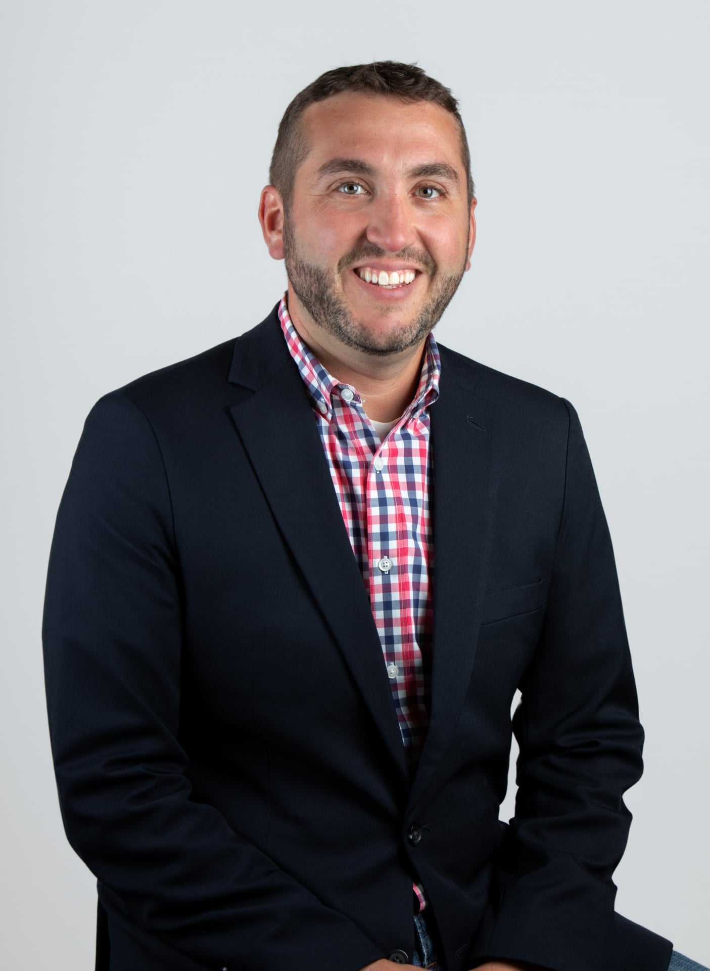 Smiling man with short hair and trimmed beard wearing a navy blazer and a red-and-blue checkered shirt, photographed in front of a light grey backdrop.