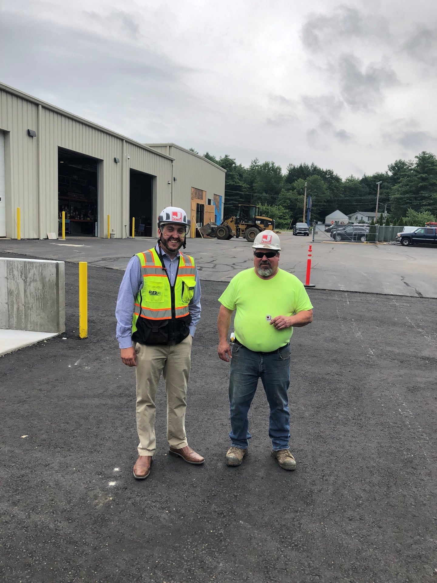 Two construction workers in hard hats standing outside a warehouse; one is wearing a bright yellow shirt and holding a safety recognition coin, while the other wears a Jewett vest and smiles at the camera.