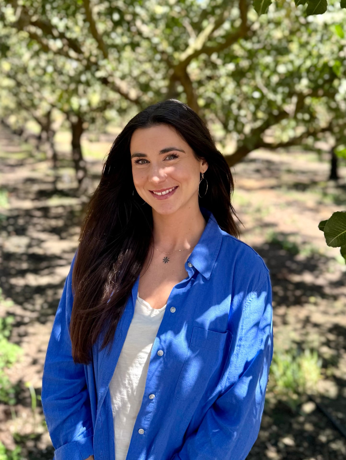 A single adult in a blue shirt stands near the orchard edge with filtered sunlight on leaves. The approachable expression and natural setting reflect the team’s responsive partner support—from scheduling to communication with growers and buyers.