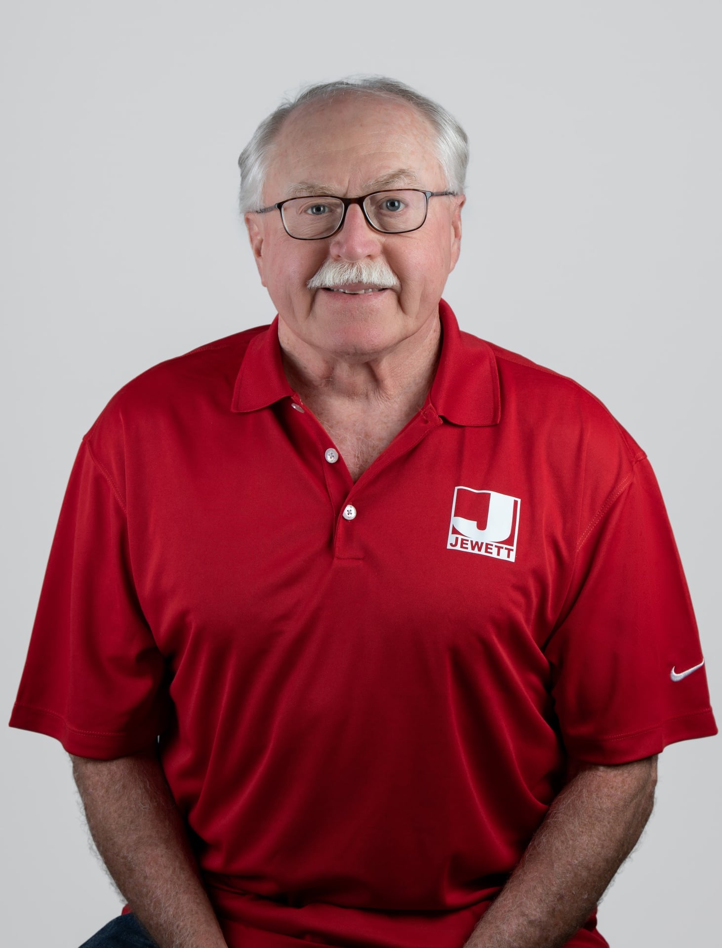 George Holcomb, wearing a red Jewett-branded polo, sits for a professional portrait against a light gray background.