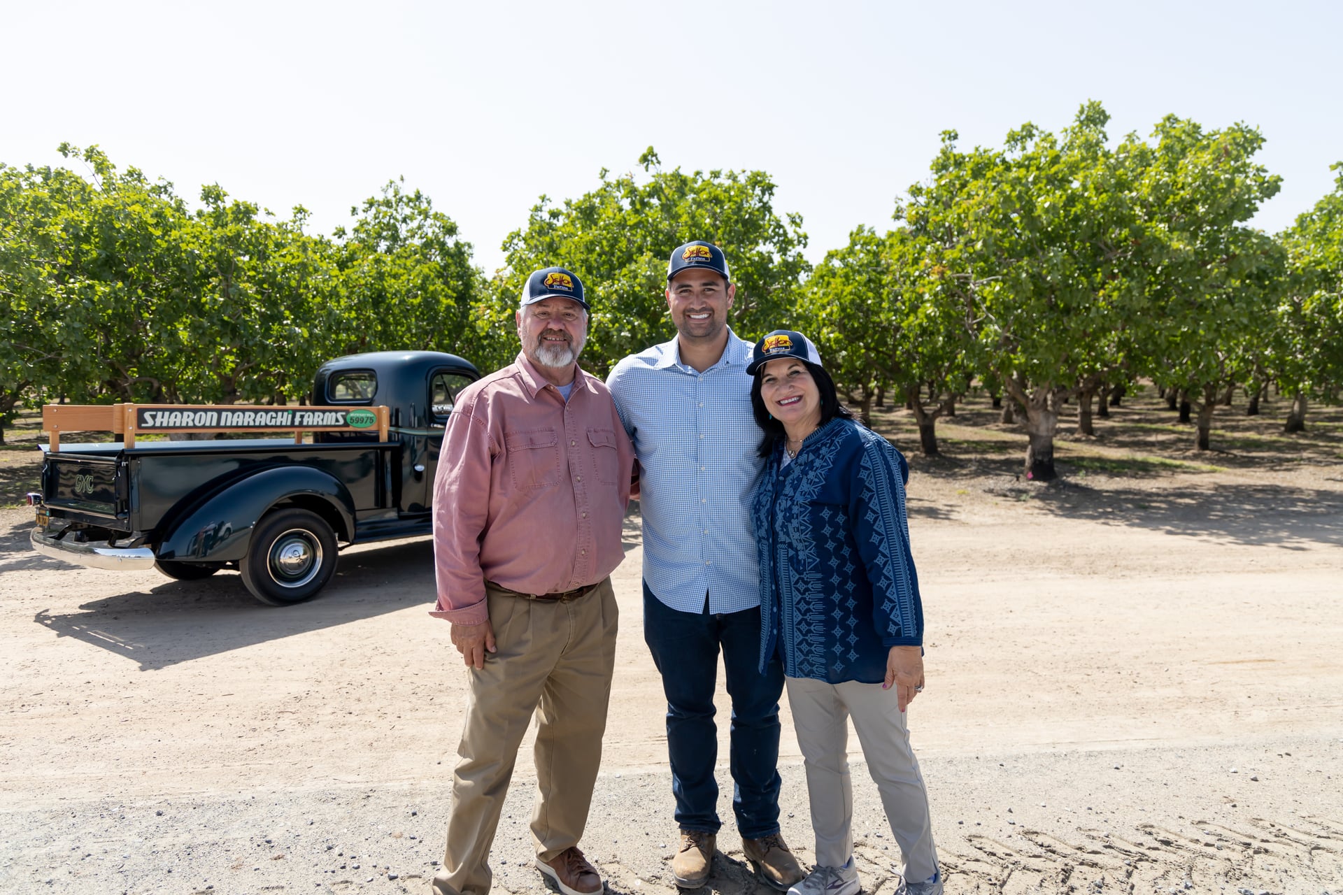 Three team members stand on an orchard lane near a vintage farm truck. Sunlit trees frame the group, illustrating Sharon Naraghi Farms’ people-first approach—relationship-driven service for growers, handlers, and buyers alongside premium crop production.
